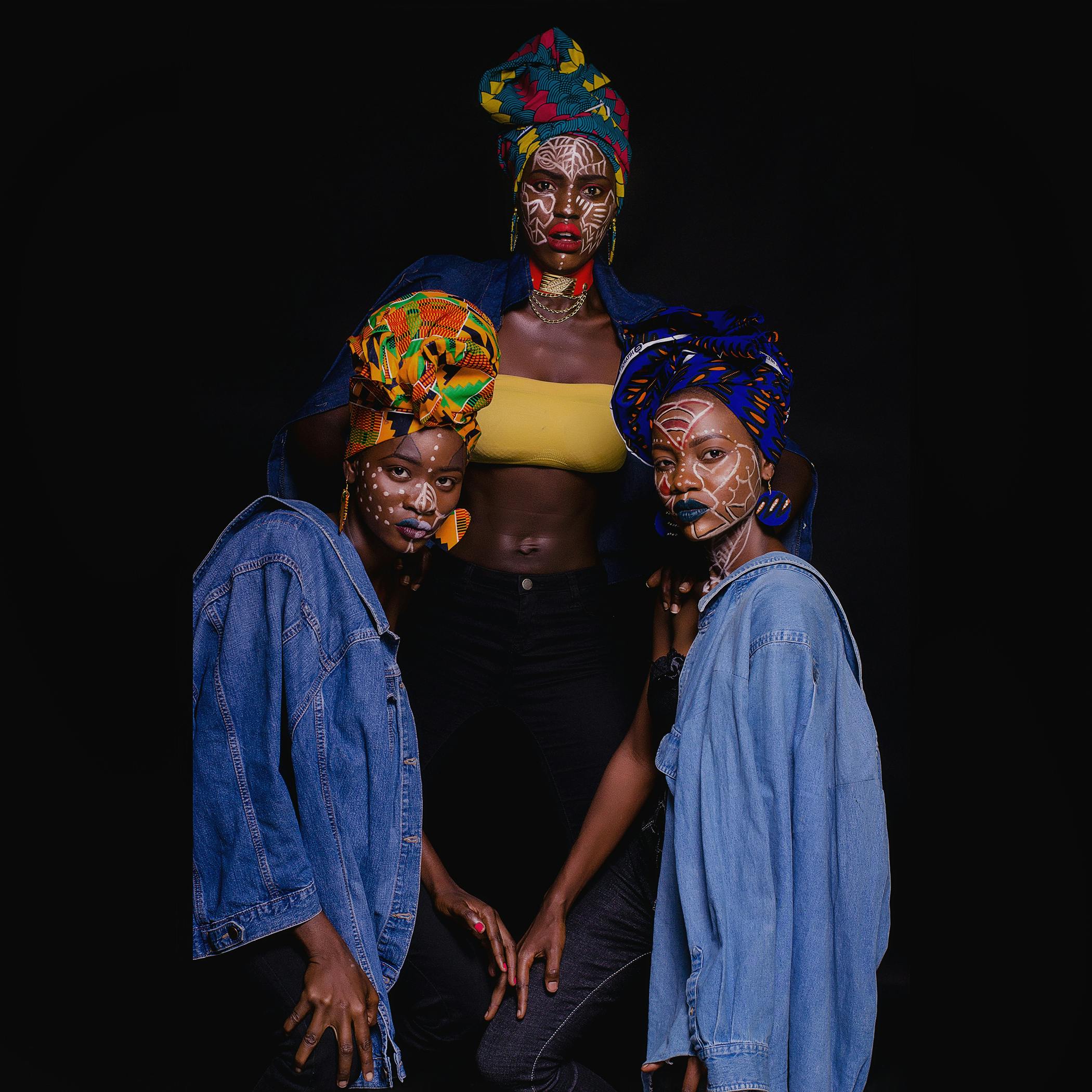 Captivating portrait of three African women in colorful headscarves and denim, adorned with face paint.
